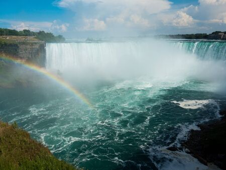 Beautiful rainbow on sunny day over Niagara Falls pool with Horseshow Falls in background. No people or ships visible. Blue sky and clouds over falls.の写真素材