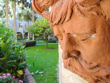 Closeup of beautiful terracotta face wall decor and gardens with mansion in background. Detailed shot of old mans bearded face in front of flowers and grass.の写真素材