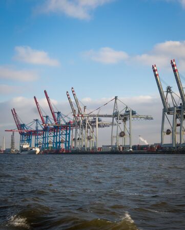 Cranes at Hamburg port cargo docks unloading freight ship containers on sunny day. No people visible, Elbe river in front. Copyspace availableの写真素材