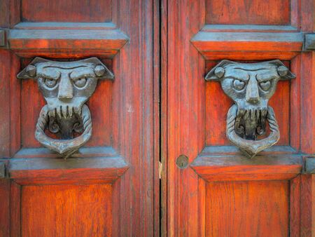 Closeup of two old and worn italian doorknockers shaped like demons or devils on red wooden door....