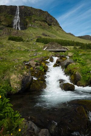 Beautiful Bjarnarfoss waterfall atop green meadows on Snaefellsnes peninsula in island. River flowing down the hills. Sunny summer day with clear blue sky, afternoon, no people visible.の写真素材