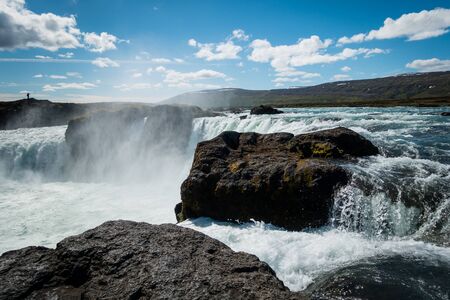Mighty Godafoss waterfall near Akureyri Iceland on beautiful sunny day with mist hanging in air. Blue sky with a few clouds. Silhouette of single person standing in distance.の写真素材