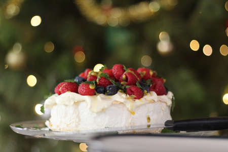Pavlova with cream & berries, Australian dessert. In the background is the Christmas tree with fairy lights and baubles.の写真素材