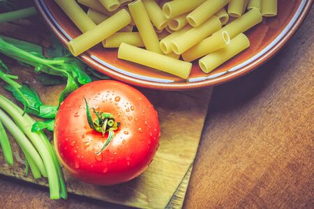 Fresh tomato next to plate full of raw uncooked macaroni. Top view with copy spaceの写真素材