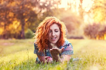 Girl with red curly hair and blue floral dress lying on her stomach in the park and looking at the palm of her hand. Plenty of copy spaceの写真素材