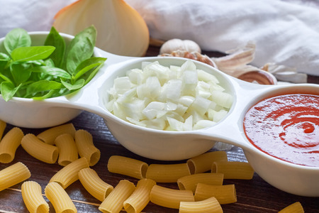 Basil, sliced onions and tomato paste in white bowl with raw pasta and garlic scattered around on wooden background. Concept image for Italian cuisineの写真素材