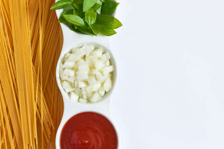 Basil, sliced onions and tomato paste in white bowl with raw spaghetti scattered around on white...