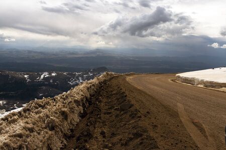 Dirt road on the summit of a mountain covered in snow with a valley of lush green forest in the...