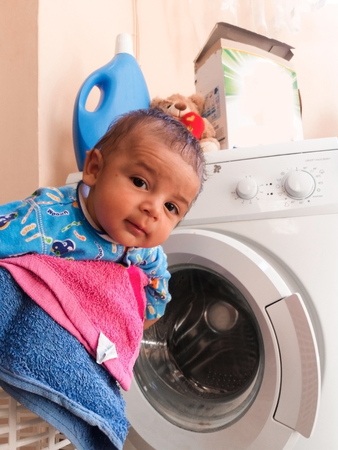 tree months old baby boy in clothes basket with washing machine and detergents in background, hellping mum to wash his clothesの写真素材