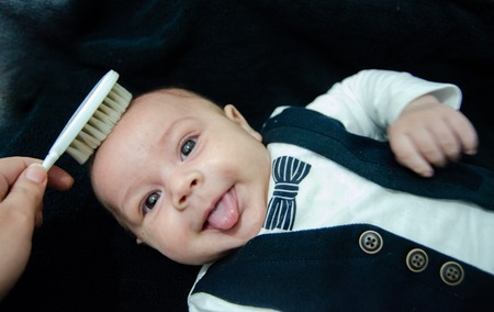 Mother brushing hair of tree months old newborn baby boy dressed like businessman with bow tie, face expresion, laying on white baby changerの写真素材