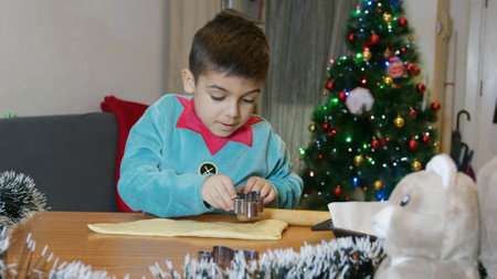 Adorable boy dressed as an elf using cookie cutters to cut gingerbread man dough. Christmas tree in the back. High quality photoの写真素材
