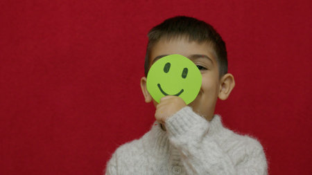 Cute child smiling and expressing his happiness with green happy emoji. Isolated on red background,. High quality photoの写真素材