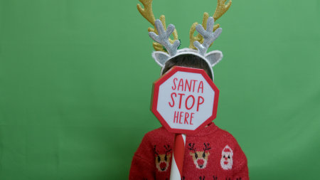 Adorable kid with double headband christmas deer antlers showing sign santa stop here. Isolated on green. High quality photoの写真素材
