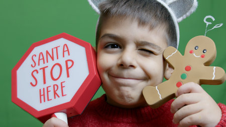 Cute boy with christmas sweater and deer alntlers holding sign Santa stop here and gingerbread man. Isolated on green background,. High quality photoの写真素材