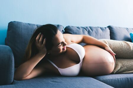 happy pregnant woman showing belly lying on gray sofaの写真素材