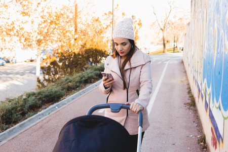 Mother with cell phone and baby carriage walking in parkの写真素材