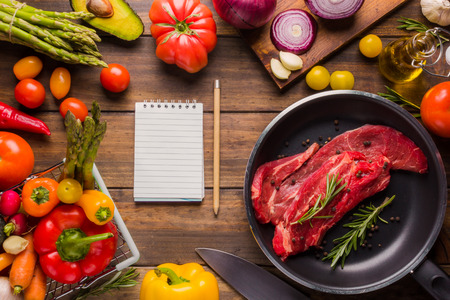 Meat seasoned and ready to cook in a pan on a wooden board and ingredients around it.の写真素材