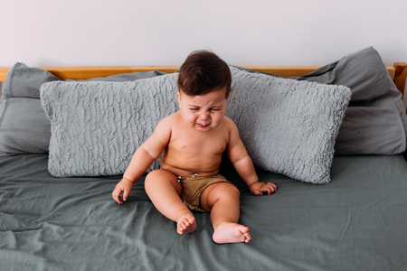 Annoyed baby crying sitting on the bed in the room wearing shorts, frustration conceptの写真素材