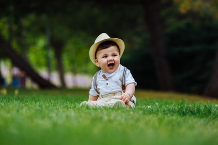 Funny and chubby baby sitting on the grass dressed in a vintage look of hat and suspenders, enjoying a spring day in the parkの写真素材
