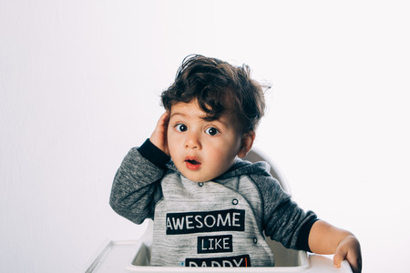 little 1 year old boy sitting with a very funny expression on his face, on a white background