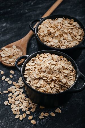 Top view of two black bowls and spoon with raw oatmeal on a black background. Breakfast with fibers for a balanced diet. Oat photoの写真素材