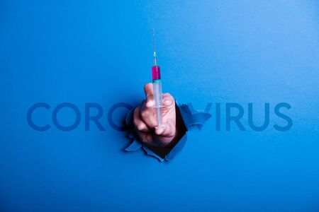 Human hand breaking a blue paper, in the hand is a syringe with the medicine against the coronavirus, concept in the fight against the coronavirusの写真素材