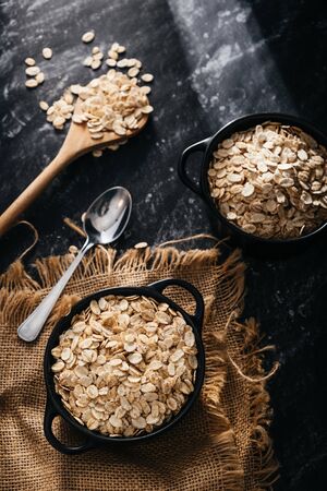 Top view of two black bowls and spoon with raw oatmeal on a black background. Breakfast with fibers for a balanced diet. Oat photoの写真素材