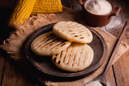 Arepas, South American food made with corn flour. Three arepas on a wooden plate and next to it a spoon and bowl full of cornmeal on a wooden backgroundの写真素材