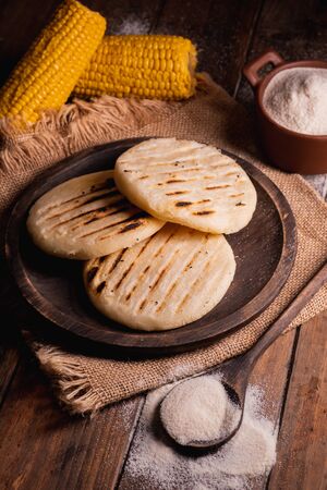 Arepas, South American food made with corn flour. Three arepas on a wooden plate and next to it a spoon and bowl full of cornmeal on a wooden backgroundの写真素材