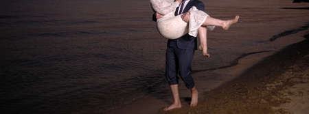 Happy bride and groom walk barefoot by the beach on sunsetの写真素材