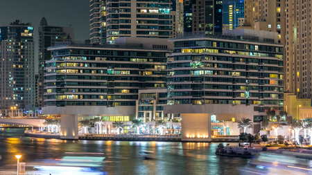 Promenade in Dubai Marina timelapse at night, UAE. View from bridge with palms, floating boats and modern towers. Dubai Marina is a district in Dubai with artificial canal city who accommodates more than 120,000 people at Persian Gulf.の写真素材