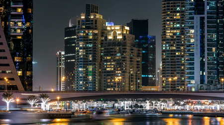 Promenade and bridge with traffic over canal in Dubai Marina timelapse at night, UAE. View from embankment  with palms, boats and towers. Dubai Marina is a district in Dubai with artificial canal city who accommodates more than 120,000 people at Gulf.の写真素材