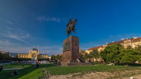 Monument of the Croatian King Tomislav timelapse hyperlapse and art pavilion in colorful park, in Zagreb, capital of Croatia. Blue cloudy sky before sunsetの写真素材