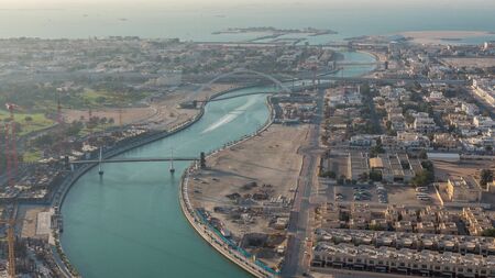 Dubai water canal with footbridge during sunset aerial timelapse from Downtown skyscrapers rooftop. Floating boats and construction site with cranesの写真素材