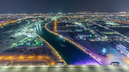 Dubai water canal with footbridge aerial night timelapse from Downtown skyscrapers rooftop. Illuminated waterfront. Floating boats and construction site with cranesの写真素材