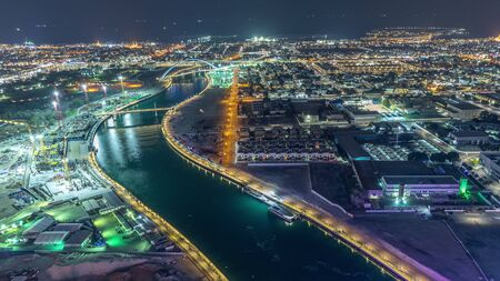 Dubai water canal with footbridge aerial night timelapse from Downtown skyscrapers rooftop. Illuminated waterfront. Floating boats and construction site with cranesの写真素材