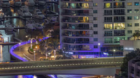Many yachts and boats are parked in harbor behind skyscraper with glowing windows aerial night...