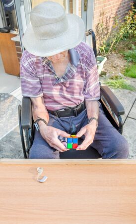 Elderly man seated in a wheelchair tries to work out a cube puzzle as therapy for his diminished mental capacity due to brain damage and stroke caused by a serious fall and injury at his home.の写真素材