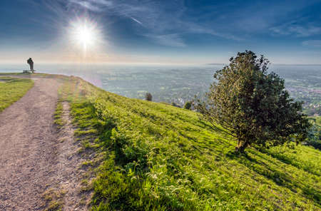 A distant human figure,standing, watching in awe the spectacle of the summer sun rising over the Worcestershire landscape, seen from the summit of North Hill, Malvern.の写真素材