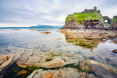 Next to the peaceful waters at low tide,,in mid summer,remains of an acient,historic Scottish castle,overlook,smooth surface of the Loch and many boulders and pebbles in seawater pools.の写真素材