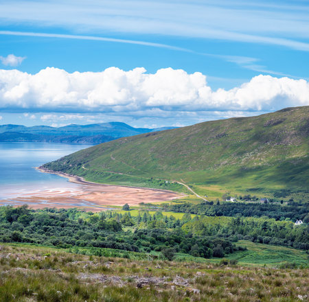 Views of the sandy bay,calm sea,blue sky and Isle of Skye in the hazy distance,green grass covered hills,line of cumulus clouds hangigng over the hills.の写真素材