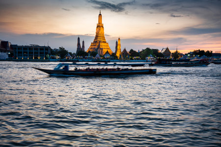 At dusk,the silhouette of a Thai Long Tail boat drifting past the golden pagodas of the temple complex,as they point towards clear sunset skies,seen from opposite side of Bangkoks main river.の写真素材
