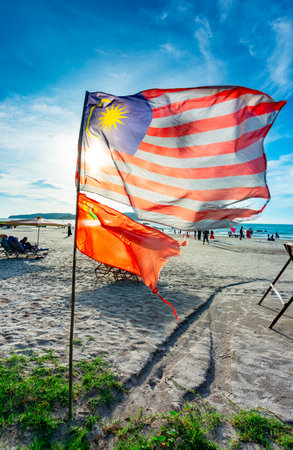 At Cenang beach ,planted into the sand,the Malaysian flag flaps in the winds coming in from the Andaman Sea,as people beyond parasail and walk along the sandy seashore.の写真素材