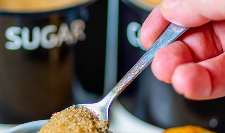Freshly poured espresso, hot and ready to drink at breakfast, a silver spoon about to mix in brown sugar to sweeten, on a wood board, a bread roll, sugar and coffee jars, out of focus in the background.の写真素材