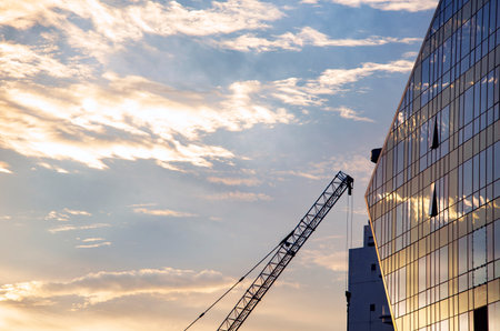 Abstract view of a high construction crane,with hook dangling,ponting towards a reflective,glass plated modern structure,mirroring the beautiful light of the surrounding city skyline.の写真素材