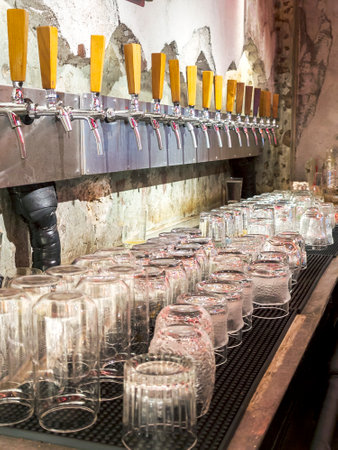 Nighttime at an small anonymous bar selling a variety of draft beers,many beer taps lined up and rows of assorted clean glasses,neatly arranged and ready for use.の写真素材