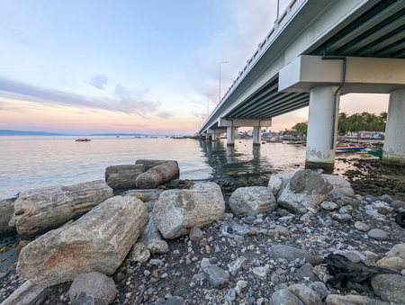 At sunset the concrete flyover takes traffic over a small stretch of water,next to fishing boats,rocks and a small beach with small huts belonging to local people.の写真素材