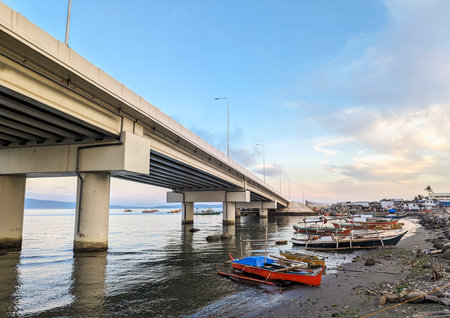 At sunset the concrete flyover takes traffic over a small stretch of water,next to fishing boats,rocks and a small beach with small huts belonging to local people.の写真素材