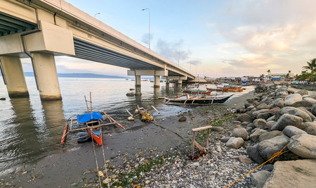 At sunset the concrete flyover takes traffic over a small stretch of water,next to fishing boats,rocks and a small beach with small huts belonging to local people.の写真素材