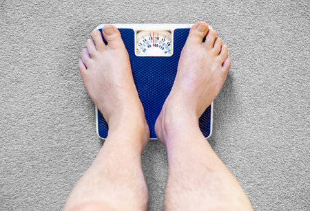 Users eye view of domestic spring scales,looking down at the bare feet and legs of a white male,to view the rotating dial,showing a readout in stones,pounds and kilograms.の写真素材